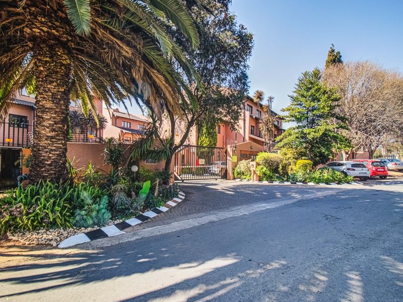 Street view of the apartment complex entrance featuring a secure gated access point, paved driveway, and established garden landscaping.