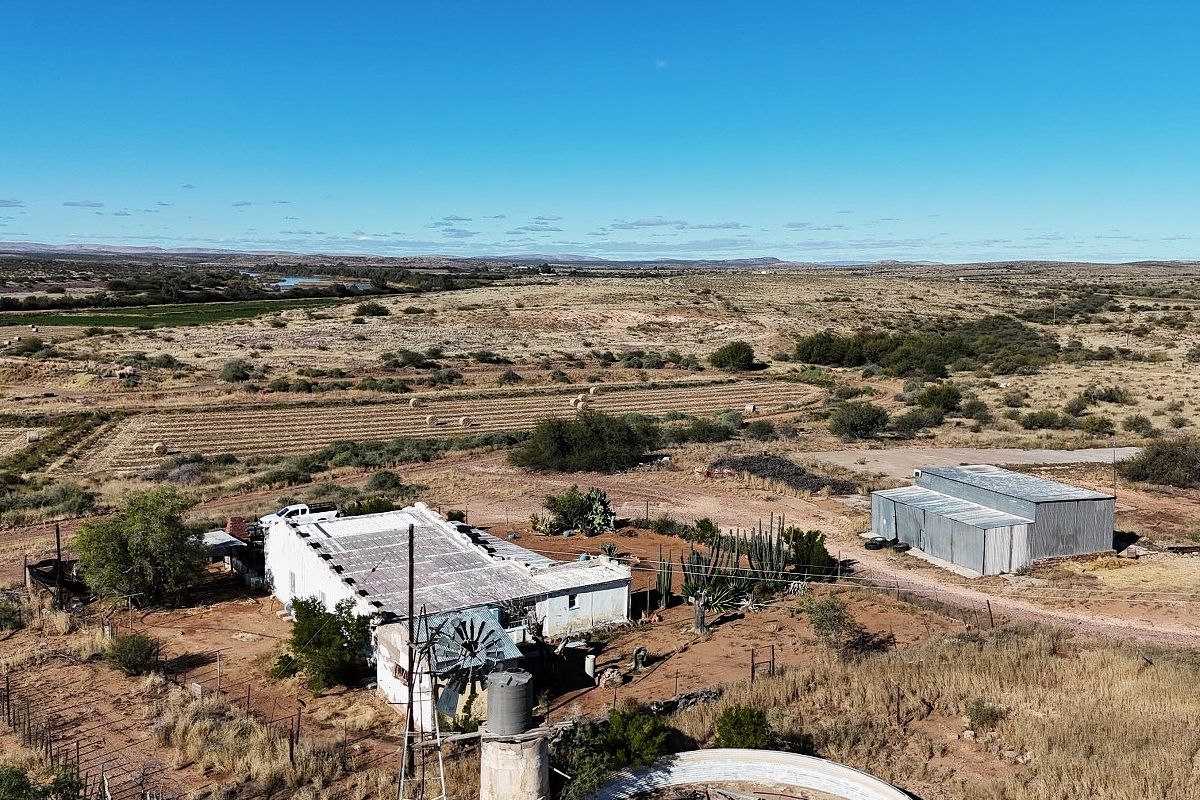 Aerial view of a rural property featuring a main dwelling, corrugated metal outbuilding, windmill, and water storage infrastructure set within a vast landscape.