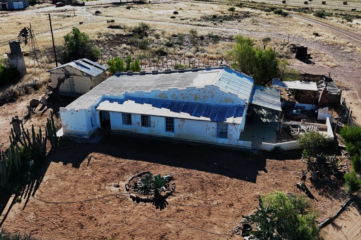 Aerial view of a rural property featuring a main dwelling with a corrugated metal roof, an adjacent outbuilding, and a wind pump on arid land.