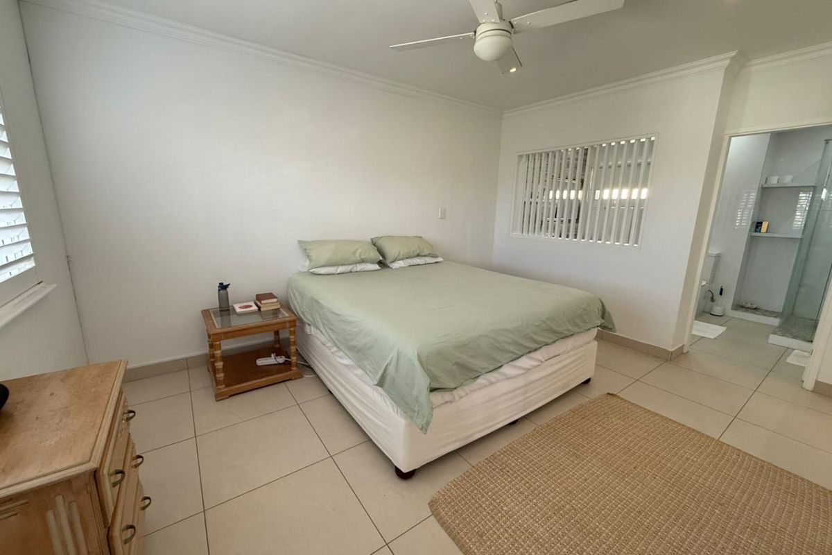 A bright bedroom featuring neutral tiled flooring, a ceiling fan, and plantation shutters, with direct access to an en-suite bathroom.