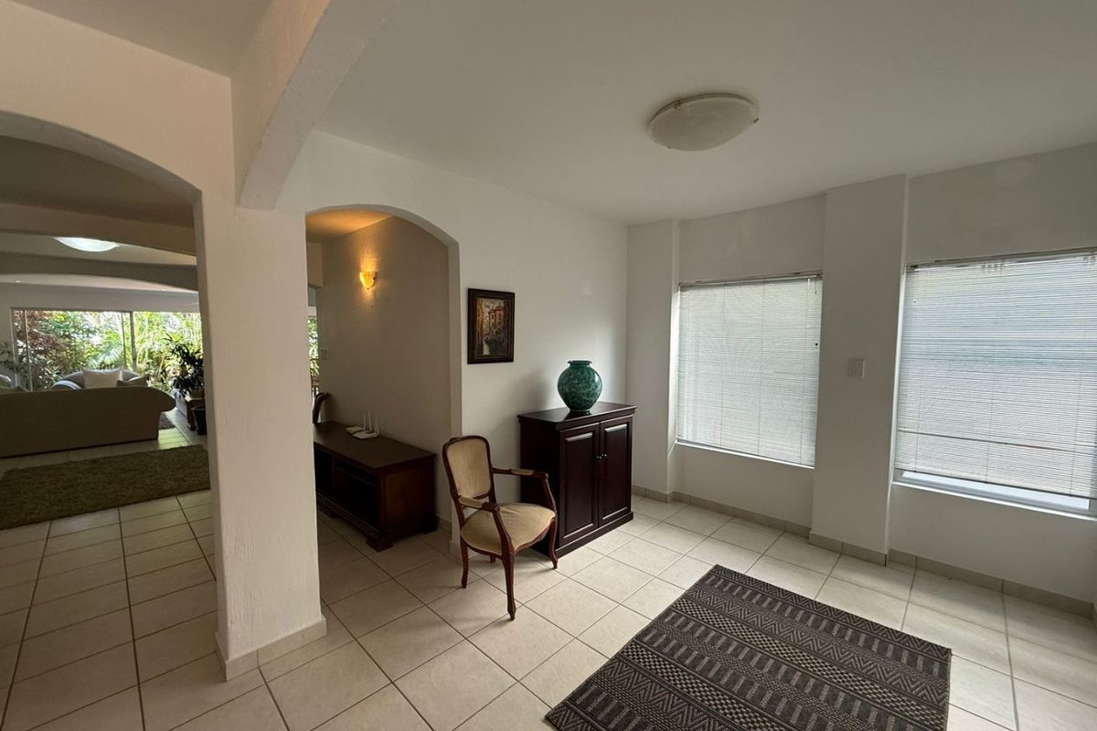 A bright living area featuring ceramic floor tiles, arched transitions between spaces, and large windows fitted with venetian blinds.