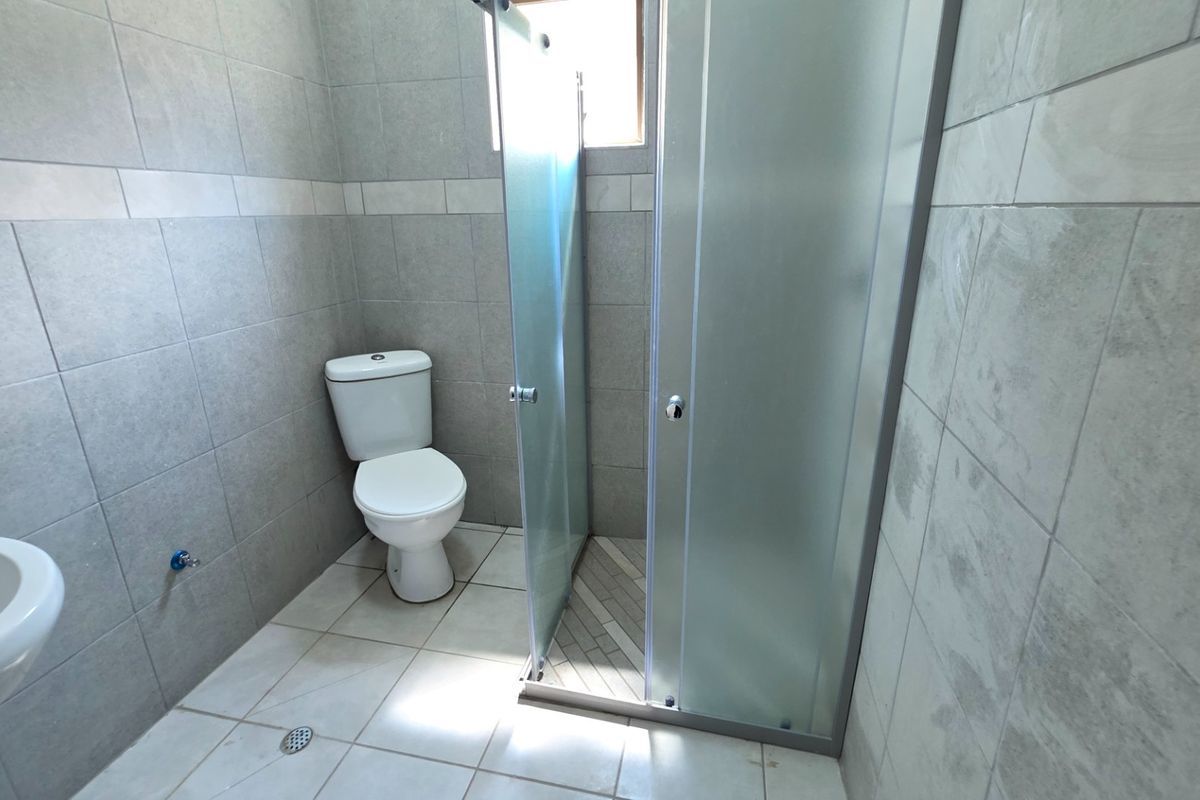 A clean, modern bathroom featuring a pedestal basin, a toilet, and a glass-enclosed shower with neutral grey wall tiles and light floor tiles.