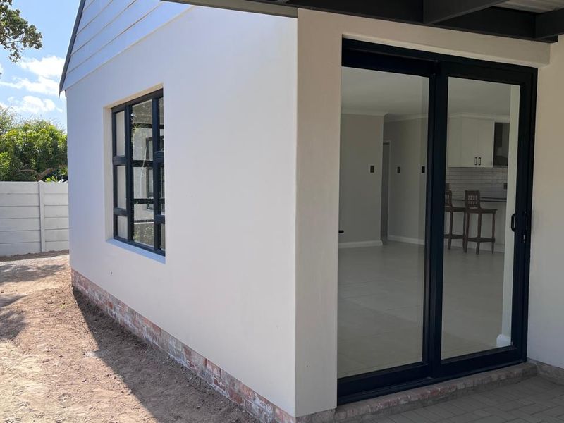 Rear exterior view showing a paved patio area, modern sliding glass doors, and aluminium window frames with a clean rendered finish.