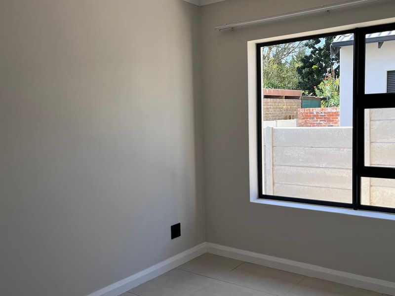 A bright, empty bedroom featuring neutral wall tones, tiled flooring, and a large window with a black aluminium frame.