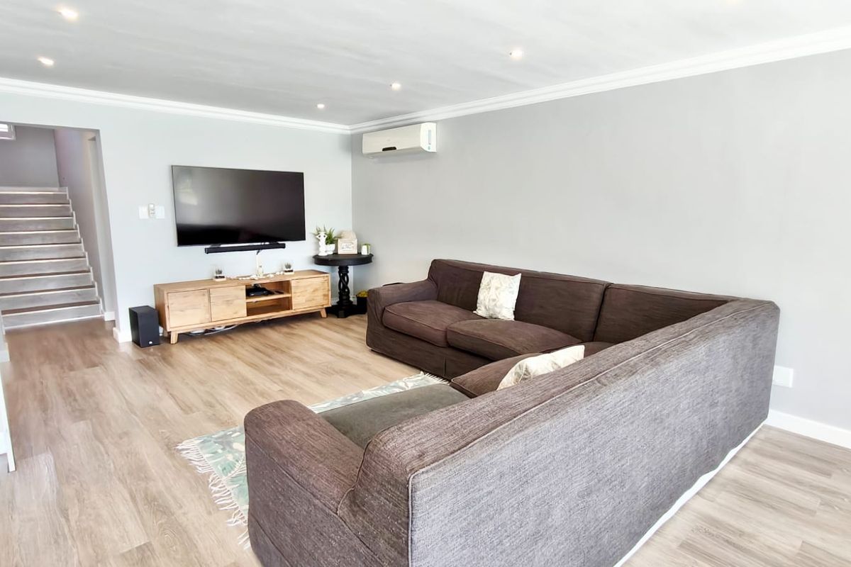 A well-maintained living room featuring laminate flooring, recessed ceiling lighting, and a wall-mounted air conditioning unit, with an adjacent staircase leading to another level.