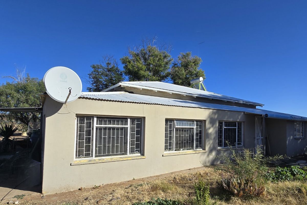 Rear view of a farm-style dwelling featuring a corrugated metal roof, solar water heating system, and security-barred windows set against a rural landscape.