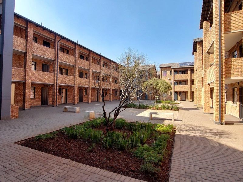A well-maintained apartment complex courtyard featuring face brick architecture, paved walkways, and landscaped garden beds under a clear sky.