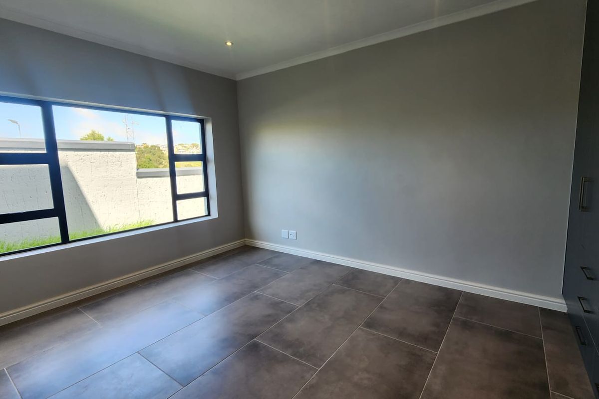 A bright, modern bedroom featuring neutral grey walls, large aluminium-framed windows, dark tiled flooring, and built-in storage cupboards.