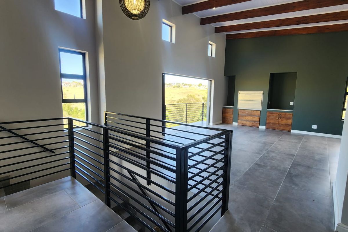 Spacious open-plan living area featuring exposed timber beams, large windows, tiled floors, and a built-in braai with direct access to a balcony.