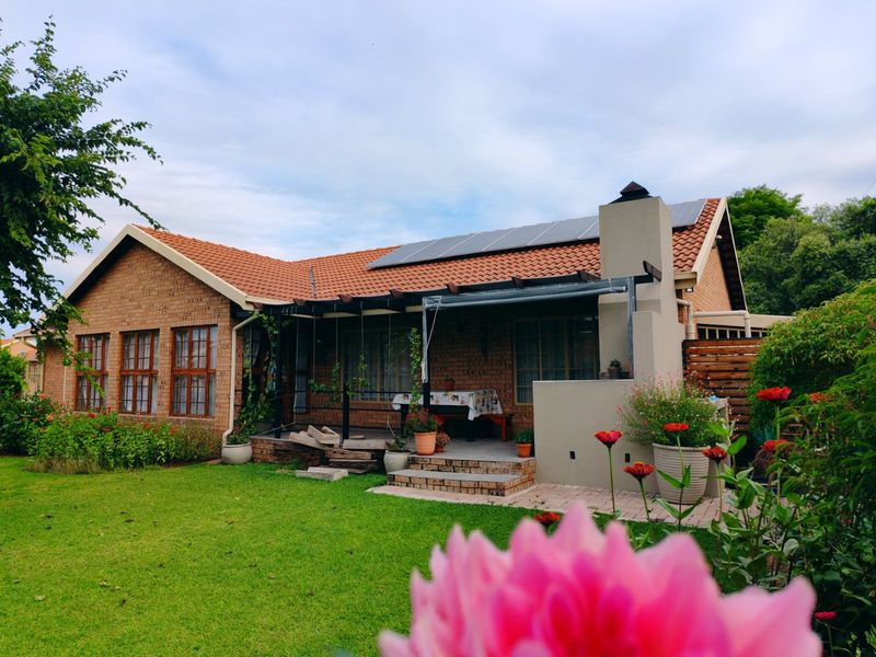 Rear view of a brick house featuring a covered patio, outdoor fireplace, and roof-mounted solar panels set within a landscaped garden.
