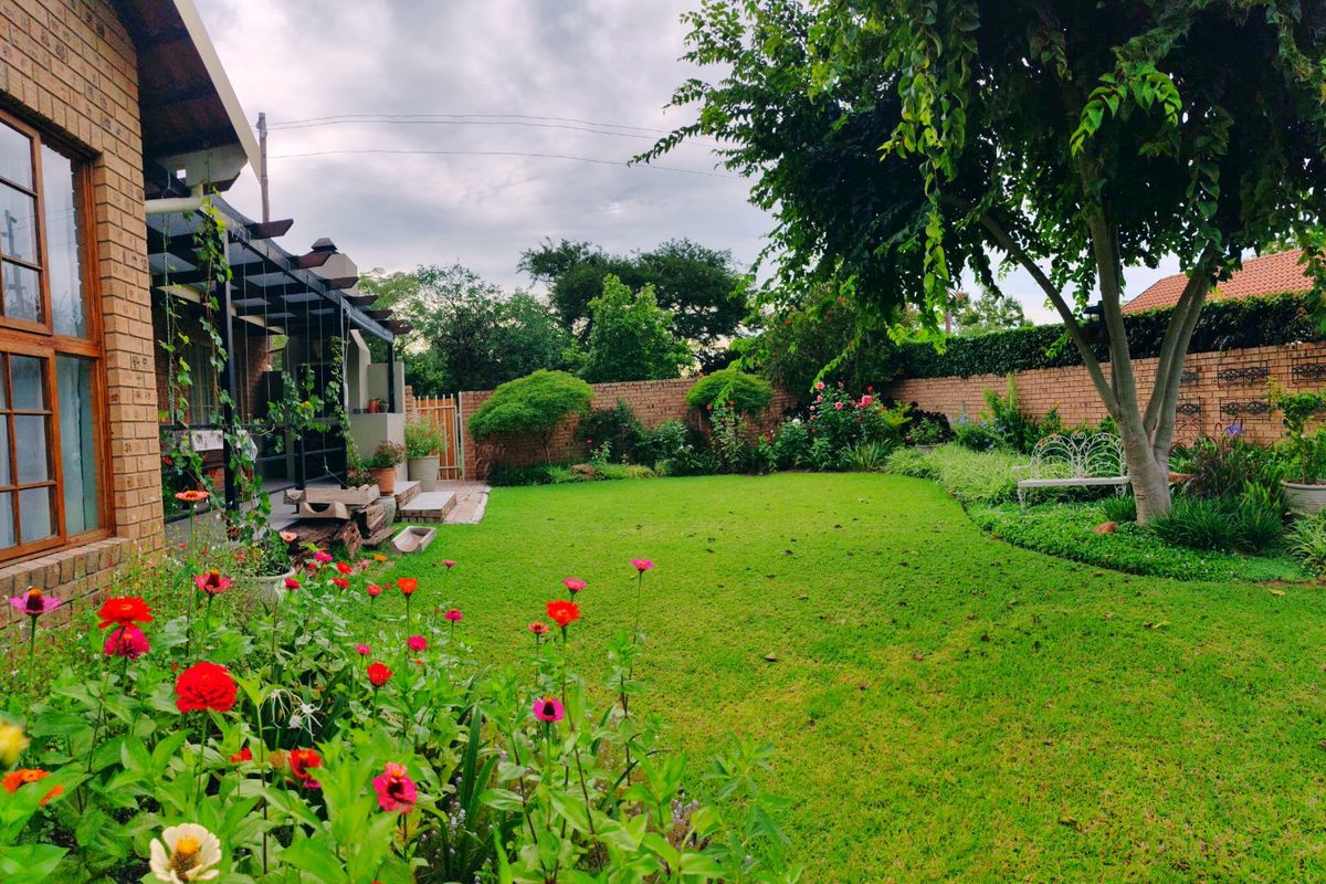 A well-maintained rear garden featuring a lush lawn, established trees, flower beds, and a covered patio area with a brick facade.