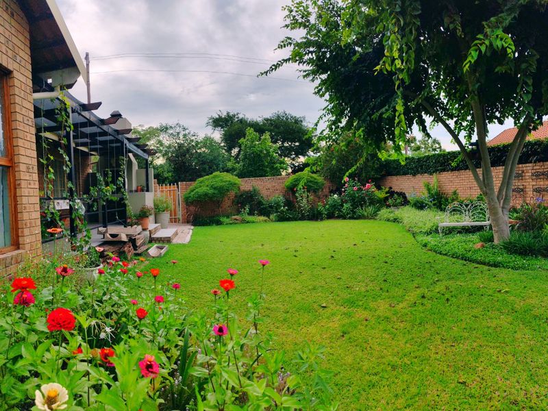 A well-maintained rear garden featuring a lush lawn, established trees, flower beds, and a covered patio area with a brick facade.