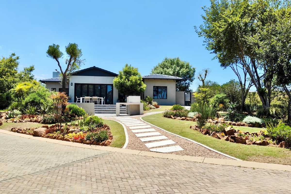 A beautifully landscaped rear garden featuring a paved driveway, stone pathway, and a raised patio area with a built-in outdoor braai.