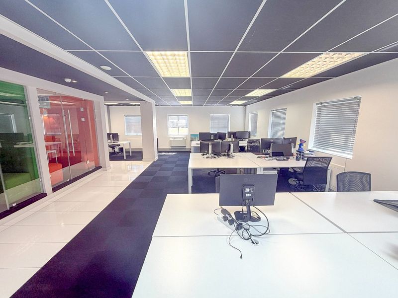 Modern open-plan office space featuring a suspended ceiling, recessed lighting, glass partitioned meeting rooms, and large windows with venetian blinds.