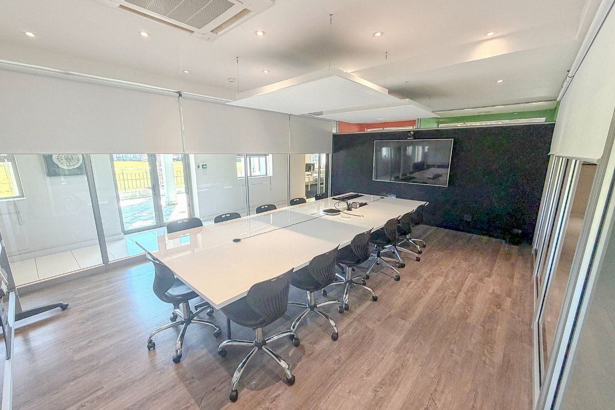 A modern, well-lit commercial boardroom featuring laminate flooring, glass partitions, integrated air conditioning, and a wall-mounted display screen.