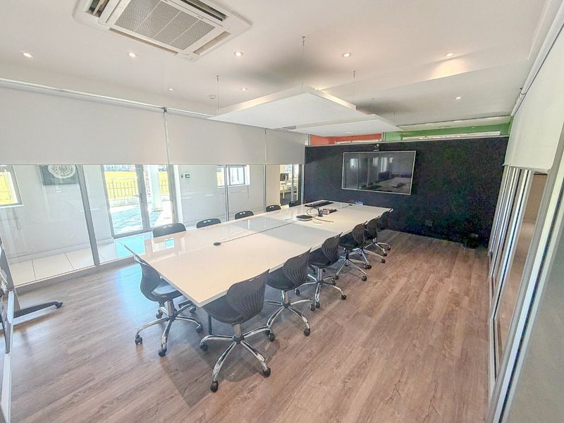 A modern, well-lit commercial boardroom featuring laminate flooring, glass partitions, integrated air conditioning, and a wall-mounted display screen.