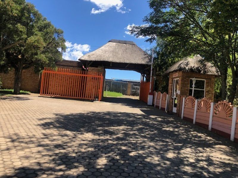 Commercial property entrance featuring a paved driveway, security guard booth, and a gated access point with a traditional thatched roof structure.