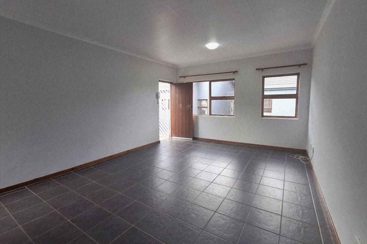 An empty living room featuring dark ceramic floor tiles, neutral walls, and natural light through wooden-framed windows and an exterior door.