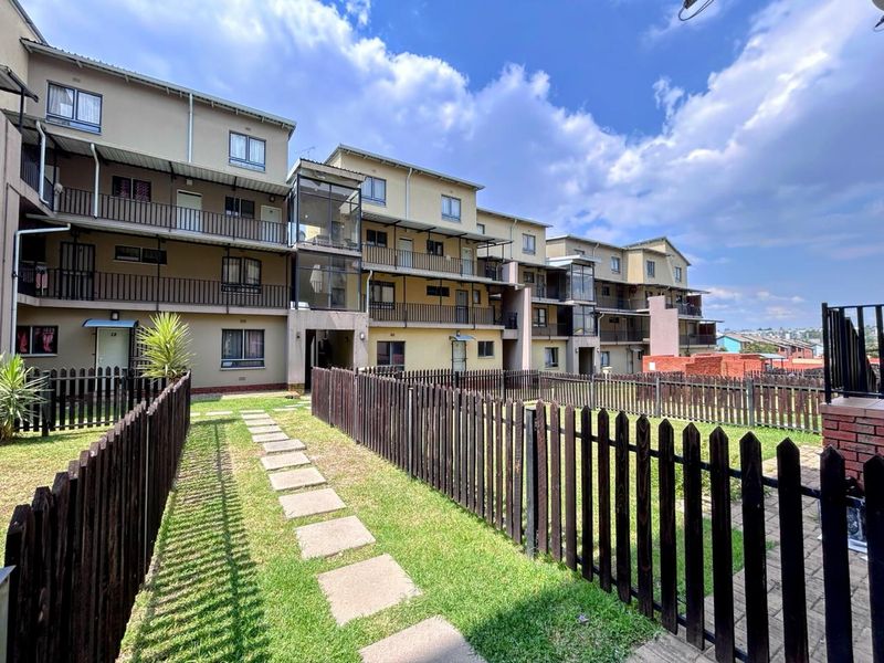 Exterior view of a multi-story apartment complex featuring private balconies, fenced garden areas, and paved walkways.
