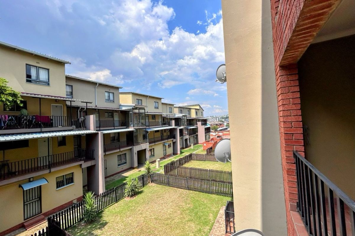 View of the apartment complex rear elevation featuring balconies, communal garden space with fencing, and brick architectural details.