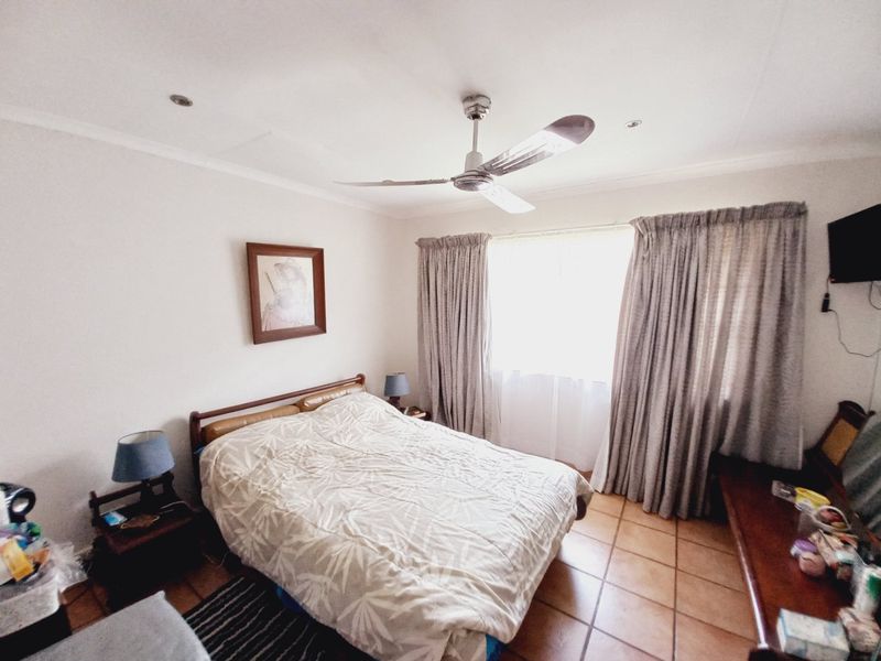 A bedroom featuring tiled flooring, a ceiling fan, and large windows with curtains, providing a functional and comfortable living space.
