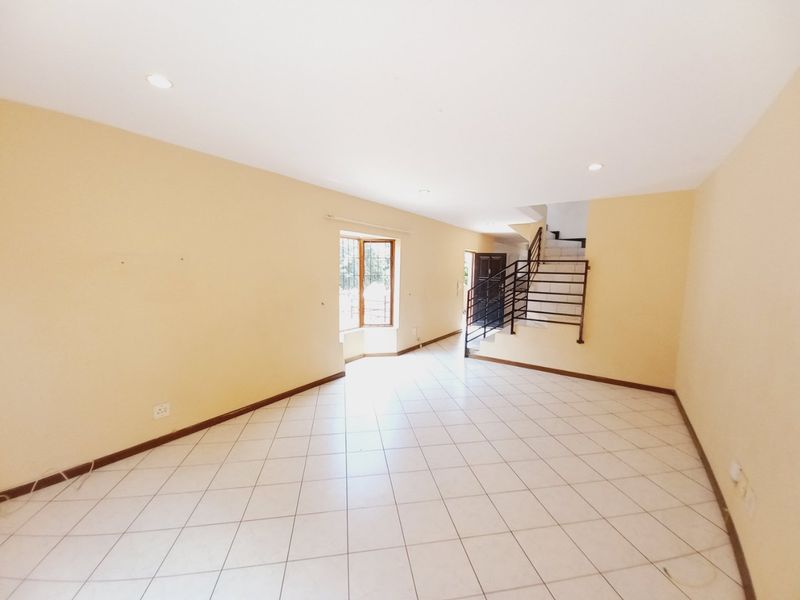An empty, open-plan living area featuring tiled flooring, a staircase with a metal railing, and a bay window, providing a bright and versatile space.