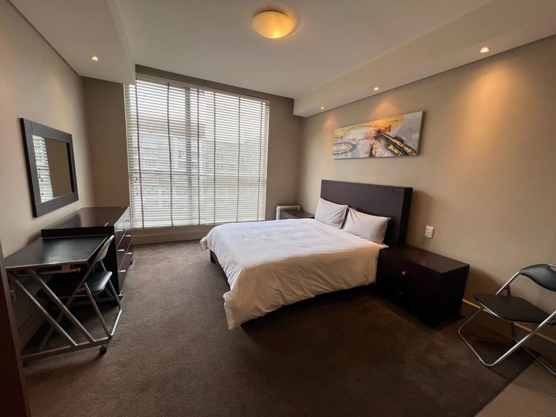 A well-maintained bedroom featuring neutral wall tones, carpeted flooring, recessed downlighting, and large windows fitted with venetian blinds.