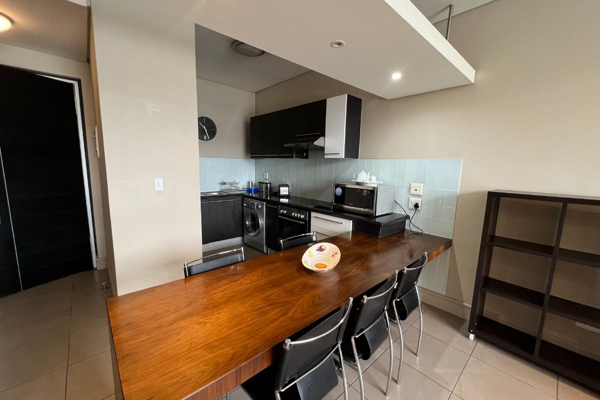 Open-plan kitchen area featuring modern cabinetry, a tiled backsplash, integrated appliances, and a built-in wooden breakfast bar, seamlessly connected to the living space.