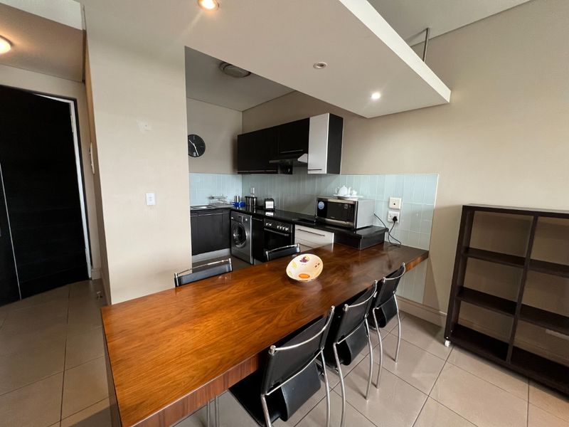 Open-plan kitchen area featuring modern cabinetry, a tiled backsplash, integrated appliances, and a built-in wooden breakfast bar, seamlessly connected to the living space.