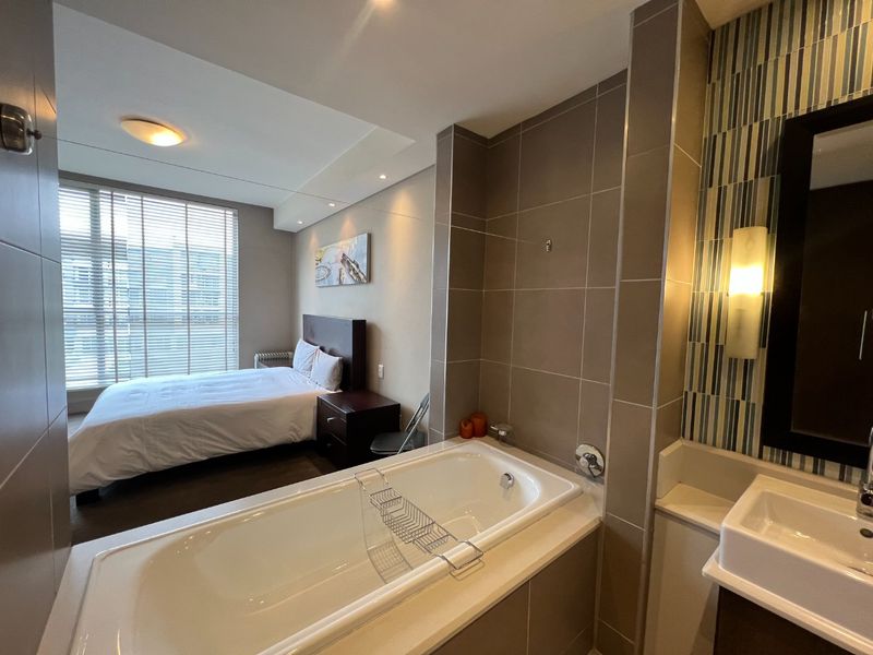 Open-plan bedroom featuring an integrated en-suite bathroom with a built-in bathtub and vanity basin, complemented by modern tiled finishes and large window blinds.