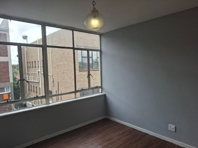 An empty bedroom featuring laminate flooring, a pendant light, and large steel-framed windows providing natural light and an urban outlook.