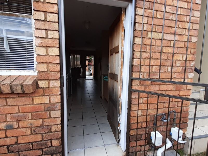 Exterior view of an apartment entrance featuring a face brick facade, security gate, and a timber door leading into a tiled interior hallway.