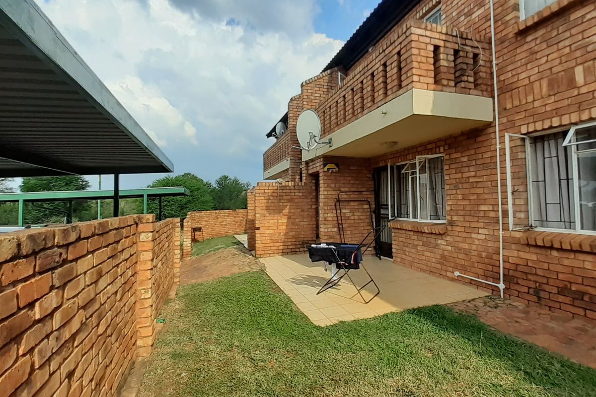 Rear view of a face-brick apartment building featuring a ground-floor tiled patio, upper-level balcony, and adjacent carport structure.