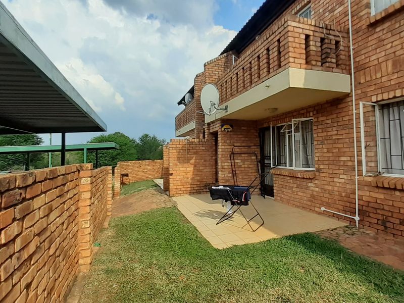 Rear view of a face-brick apartment building featuring a ground-floor tiled patio, upper-level balcony, and adjacent carport structure.
