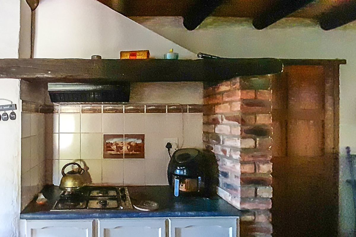 A rustic kitchen area featuring a gas hob, extractor fan, tiled backsplash, and exposed timber ceiling beams with a decorative brick pillar.