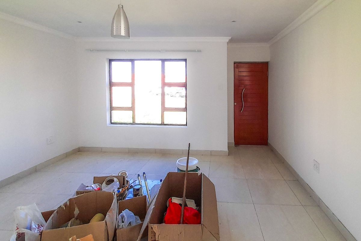 A bright, empty living room featuring neutral tiled flooring, clean white walls, a modern pendant light, and a solid timber entrance door.