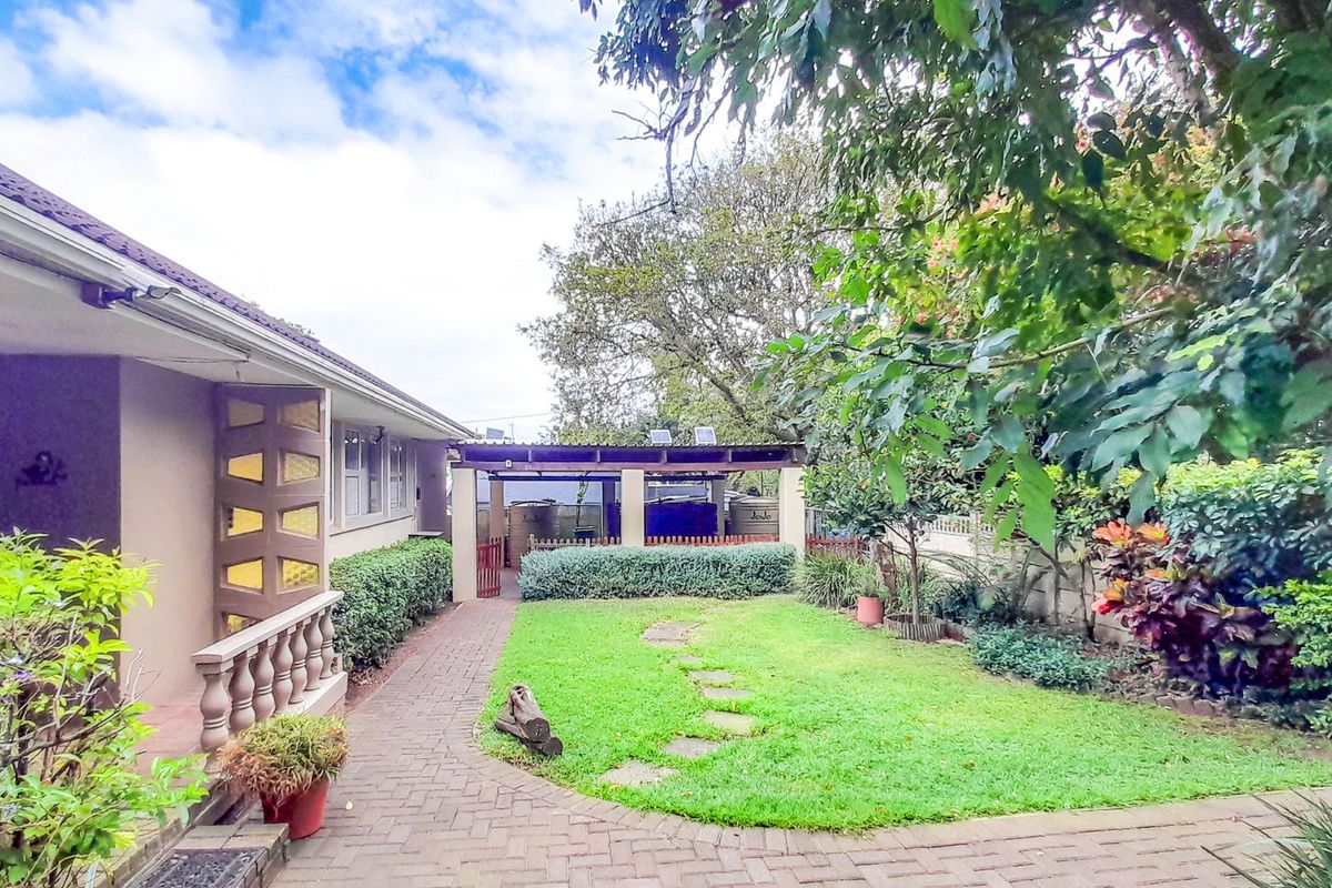 Rear garden view featuring a brick paved pathway, covered patio area, and established landscaping with water storage tanks.