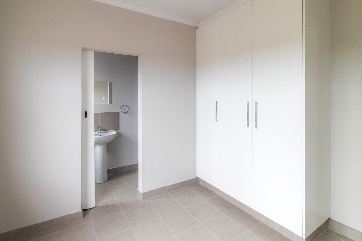 A clean, empty bedroom featuring neutral tiled flooring, ample built-in cupboard space, and an adjoining en-suite bathroom with a pedestal basin.