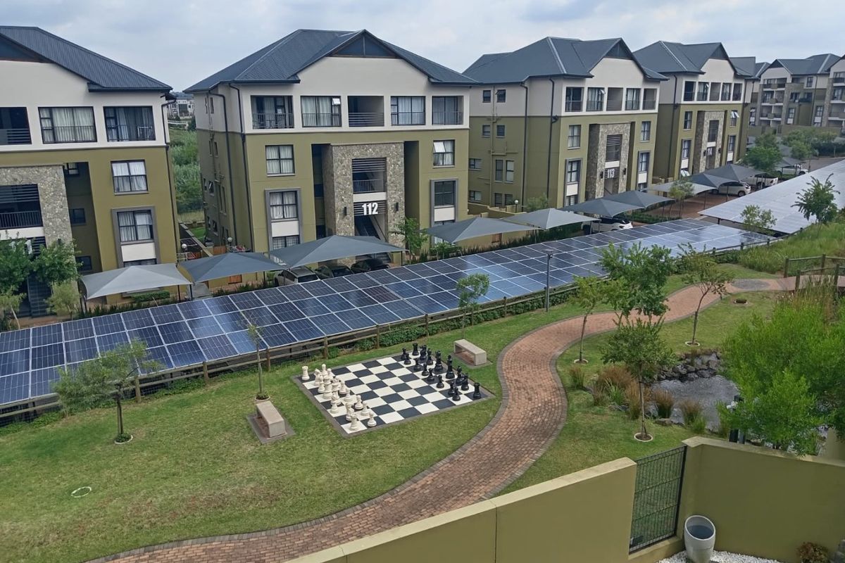 Aerial view of an apartment complex featuring landscaped gardens, a large solar panel array, paved walkways, and covered parking bays.