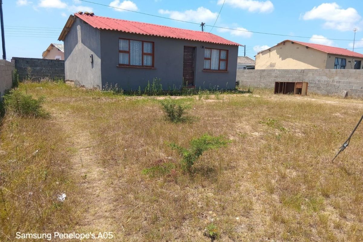 A detached house featuring a corrugated metal roof and concrete block boundary walls, set on a large, unpaved plot with overgrown vegetation.