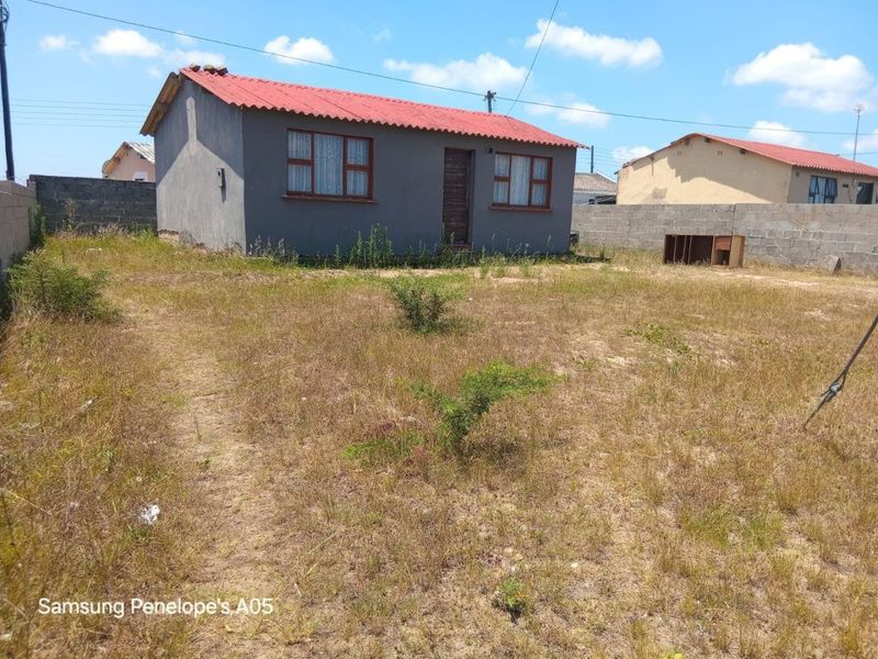 A detached house featuring a corrugated metal roof and concrete block boundary walls, set on a large, unpaved plot with overgrown vegetation.