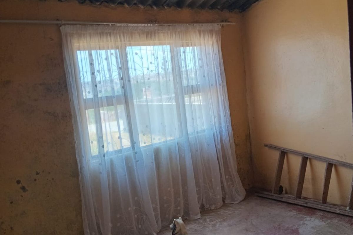A bedroom space featuring exposed corrugated iron roofing, timber rafters, and a window with security bars, requiring significant renovation and cosmetic updates.