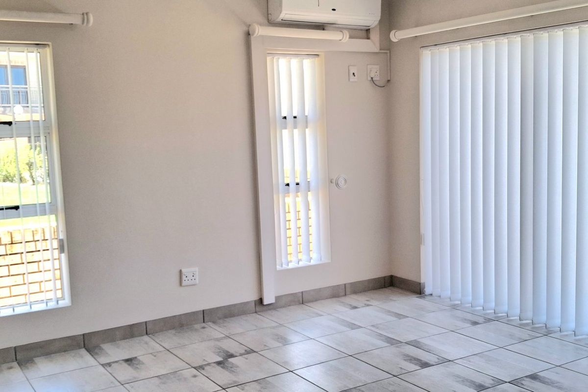 A bright, empty living area featuring neutral tiled flooring, vertical blinds, and a wall-mounted air conditioning unit.