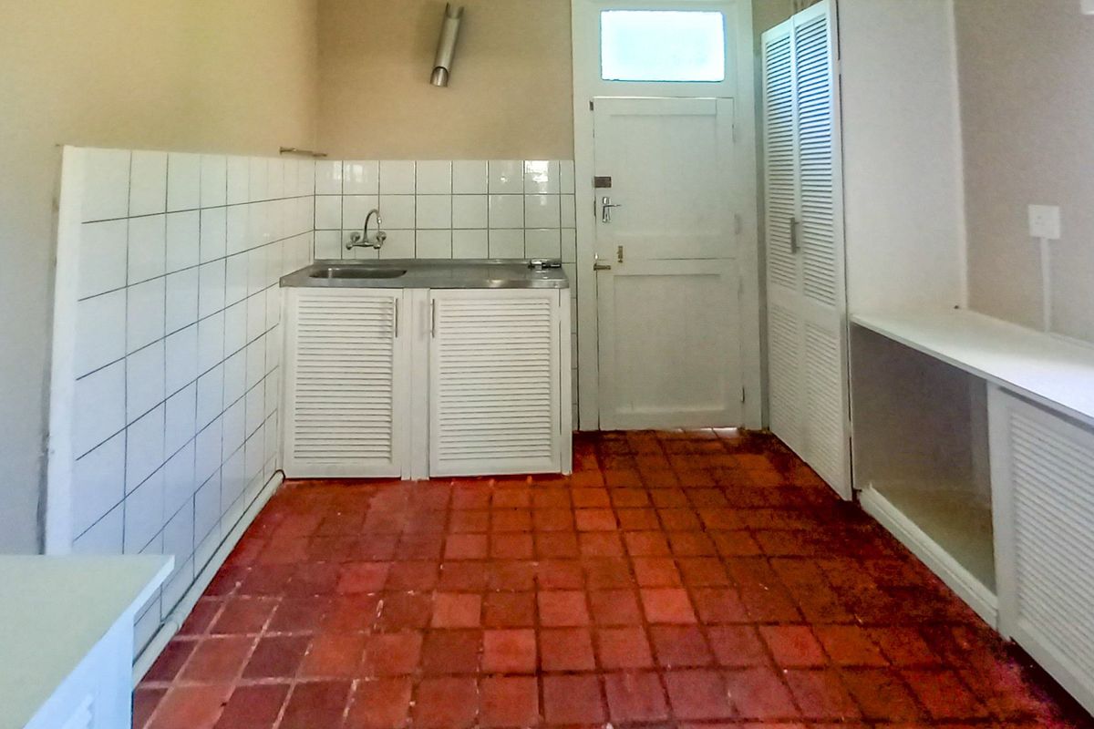 A functional, empty kitchen space featuring terracotta floor tiles, built-in cabinetry with louvered doors, and a stainless steel sink with tiled splashback.