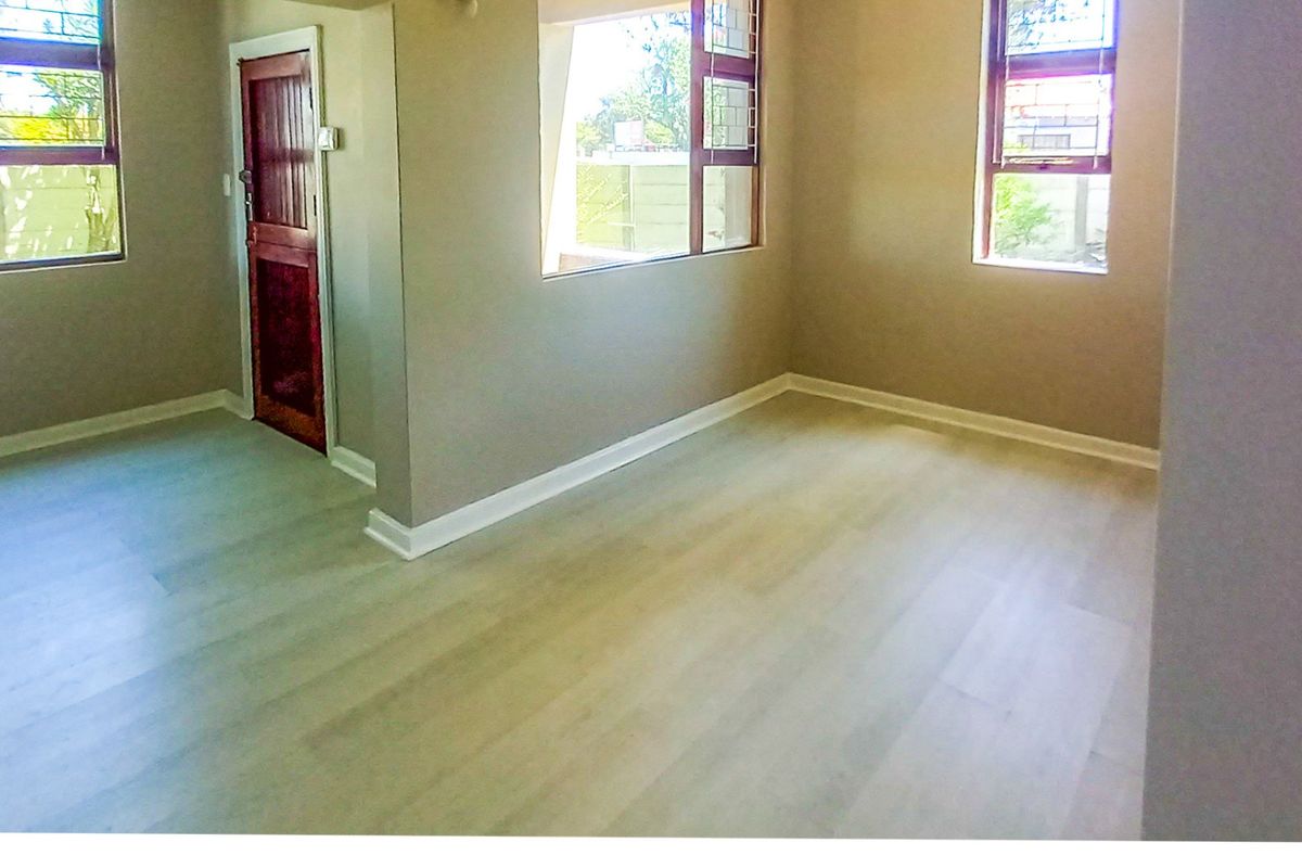 An empty living room featuring light-toned laminate flooring, neutral walls, and multiple windows with security bars, providing a bright and versatile living space.