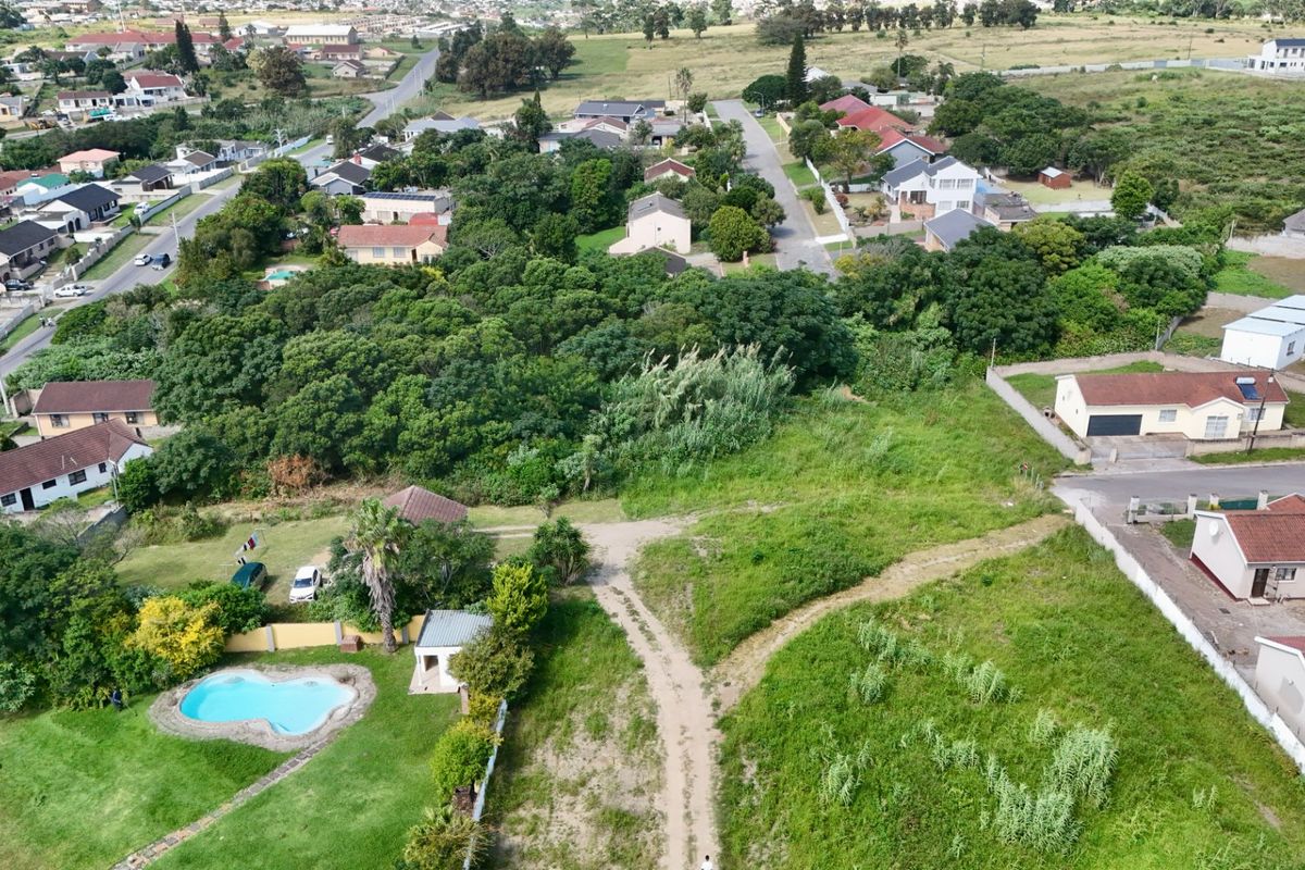 Aerial view of a vacant residential plot featuring natural vegetation, a dirt access path, and proximity to established suburban housing and a swimming pool.