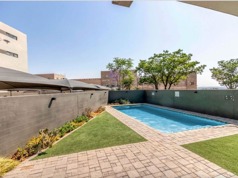 A well-maintained outdoor pool area featuring a rectangular swimming pool, paved surround, artificial lawn, and perimeter boundary walls with shade sails.