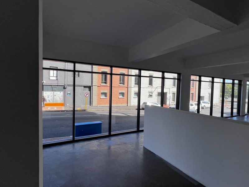 A bright, open-plan commercial office space featuring polished concrete floors, exposed structural beams, and expansive windows offering street-level views.