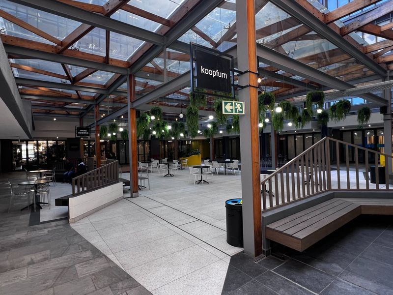 A modern commercial atrium featuring a glass roof with timber beams, stone flooring, and integrated seating areas, creating a bright and open communal space.