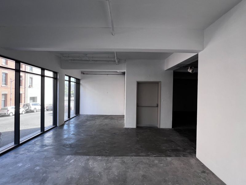 An empty, open-plan commercial space featuring large floor-to-ceiling windows, concrete flooring, and exposed ceiling infrastructure, suitable for office or retail use.
